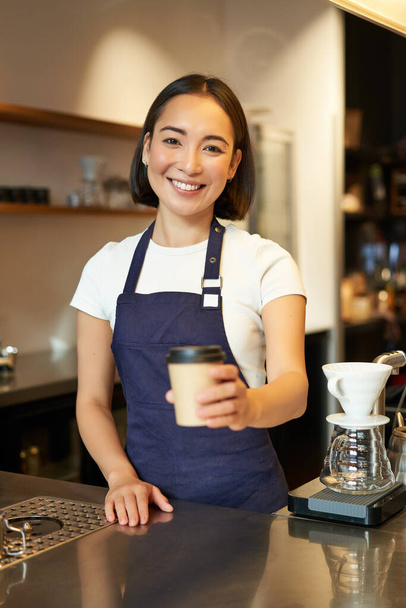 Улыбающаяся азиатка-бариста отдаёт приказы клиенту, держит чашку кофе на вынос, носит фартук, работает за прилавком в кафе. - Фото, изображение