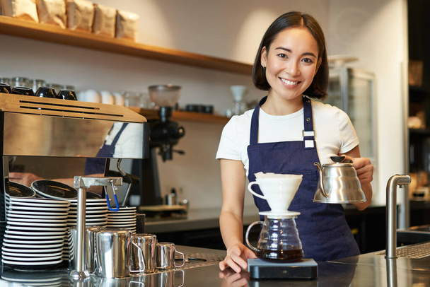 Lächelnde Barista, asiatische Barkeeperin gießt Wasser aus Wasserkocher, brüht Filterkaffee im Café hinter der Theke, trägt blaue Schürze. - Foto, Bild