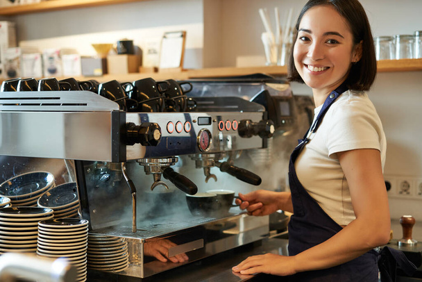 Улыбаясь симпатичной девушке, бариста, работающая с кофемашиной, готовит капучино, используя стемр на прилавке кафе. - Фото, изображение