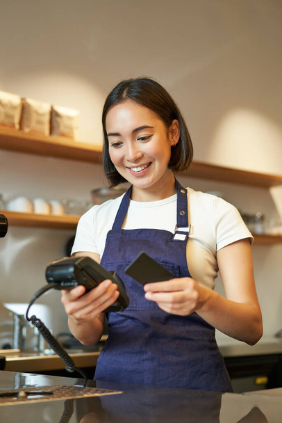 Portré mosolygós ázsiai barista, kávézó alkalmazott POS terminál és hitelkártya, segít az ügyfél fizetni érintetlen kávézóban. - Fotó, kép