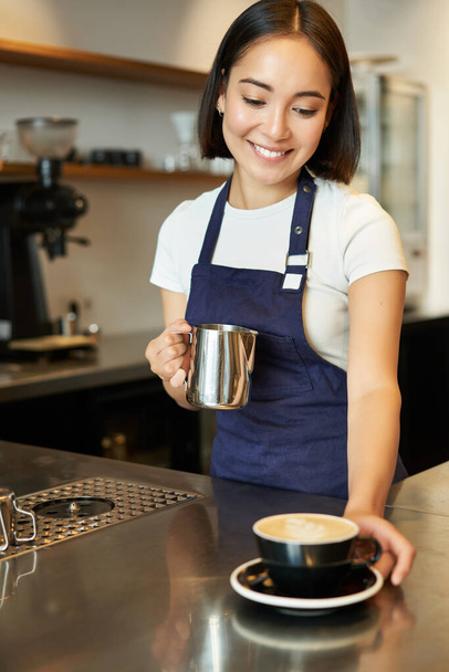 Kafede gülümseyen Asyalı bir kızın dikey fotoğrafı, kapuçino dağıtırken, sipariş verirken, kahve hazırlarken, mavi önlüklü tezgahın arkasında dururken.. - Fotoğraf, Görsel