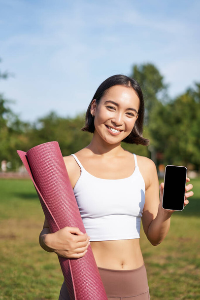 Excited fitness girl recommends application for sport and workout, shows phone screen, standing with rubber yoga mat in park after training session. - Photo, Image