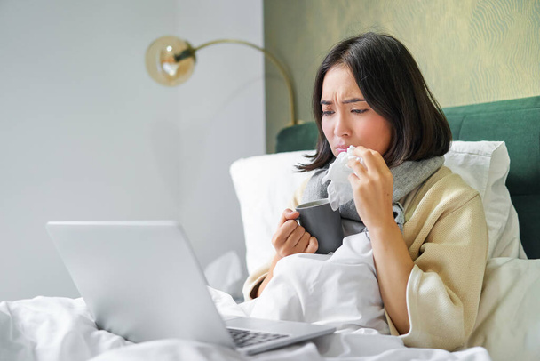 Portrait of asian girl sneezing and wiping runny nose, feeling sick, staying in bed with influenza, looking at laptop. - Foto, Bild