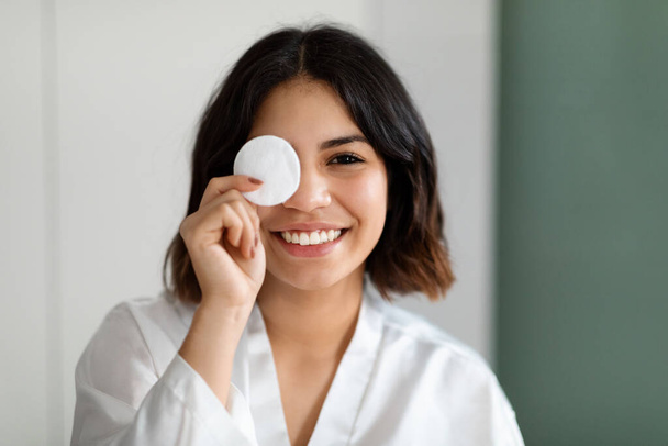 Closeup photo of cheerful happy young arabic woman wearing white silk bathrobe cover one eye with cotton pad and smiling, home interior, copy space. Face cleansing, skin care routine concept - Photo, Image