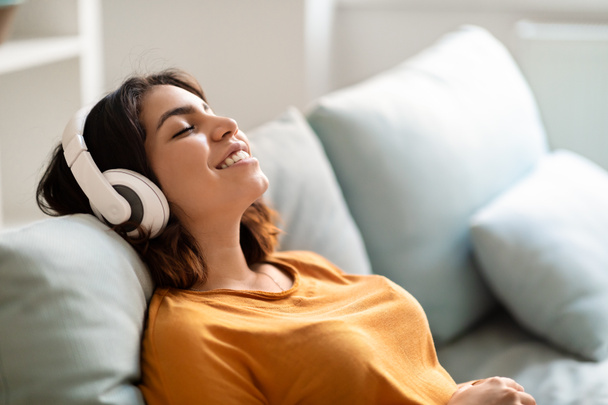 Любимое досуг. Домашняя арабская женщина слушает музыку в беспроводных наушниках, веселая ближневосточная женщина ложится на диван с закрытыми глазами, копирует пространство - Фото, изображение
