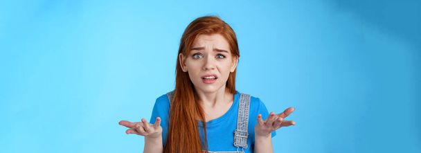 Puzzled confused redhead cute woman look ambushed, raise hands shrugging uncertain, frowning perplexed, cannot understand what happened, stand frustrated upset blue background. Copy space - Photo, Image