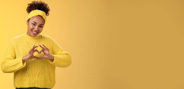 Precioso tierno elegante afro-americana chica rizado peinado diadema suéter mostrando corazón amor gesto alegremente sonriente coqueta inclinación cabeza expresar simpatía pasión deseo, posando fondo amarillo. - Foto, imagen