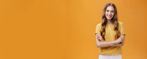 Elämäntapa. Muotokuva ystävällinen ja ystävällinen viehättävä nuori naispuolinen opiskelija rento t-paita aaltoileva luonnollinen pitkät hiukset pitämällä kädet ristissä rinnassa hymyillen laajasti ja huoleton kameran yli oranssi - Valokuva, kuva