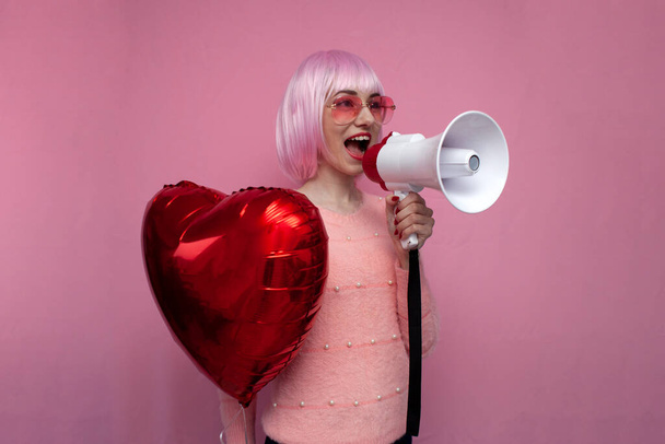 νεαρή κοπέλα με ροζ μαλλιά κρατά ένα φουσκωτό μπαλόνι καρδιά και ουρλιάζει δυνατά σε ένα ροζ φόντο, ένα σύμβολο της αγάπης, ημέρα του Αγίου Βαλεντίνου - Φωτογραφία, εικόνα