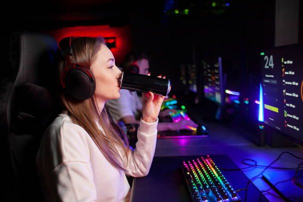 νεαρή gamer κορίτσι κάθεται σε έναν υπολογιστή και πίνει ένα ενεργειακό ποτό, μια γυναίκα παίζει σε ένα κλαμπ υπολογιστών τη νύχτα και κρατά ένα ποτό - Φωτογραφία, εικόνα