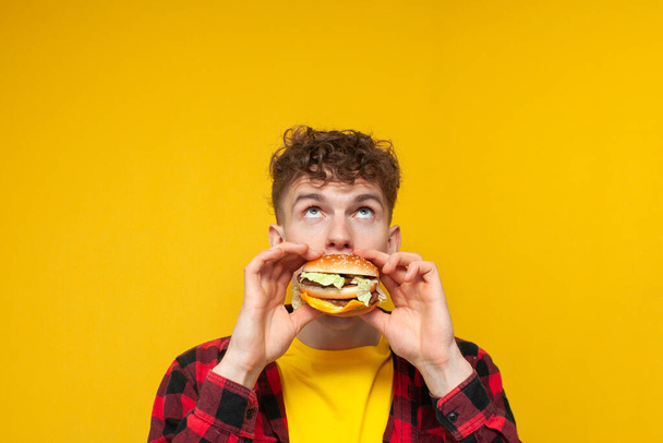 young curly guy student eats a burger on a yellow background and looks up, a man with fast food looks at the place for text - Photo, image