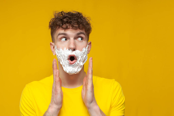 fiatal göndör fickó borotvahabbal az arcán meglepődik, és elfordul a sárga háttér, egy vicces diák borotválja a szakállát - Fotó, kép