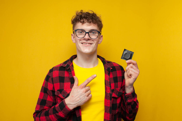 young curly guy holds a condom on a yellow background and smiles, a student shows a contraceptive, the concept of safe sex - Photo, image