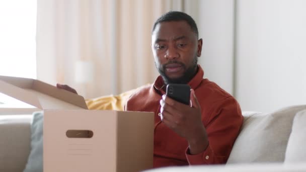 Παρακολούθηση διαδικτυακών αγορών. Νεαρός σοβαρός αφρικανός αμερικανός πελάτης ελέγχοντας την παραγγελία σε εφαρμογή smartphone και ανοίγοντας το κιβώτιο παράδοσης, ελέγχοντας την αγορά στο σπίτι, εντοπίζοντας shot, αργή κίνηση, ελεύθερος χώρος - Πλάνα, βίντεο
