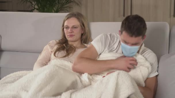CLOSE UP. Twosome watching TV covered with blanket while man with flu sneezes. Young man wears protective mask and sneezes then lady disinfects the air with spray. Winter colds and flu spread around - Imágenes, Vídeo