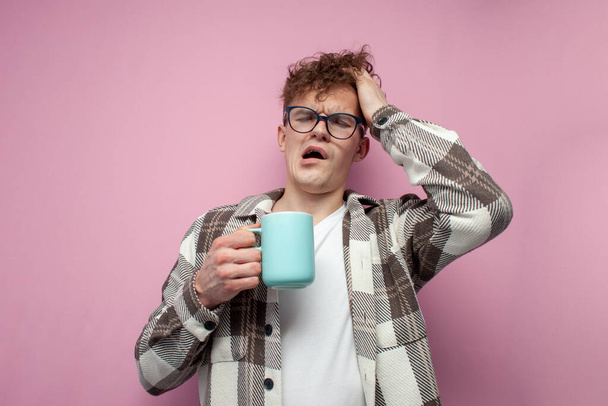 tired sleepy young guy in glasses holds a mug of coffee and yawns on a pink background, the concept of sleepiness and fatigue - Photo, Image