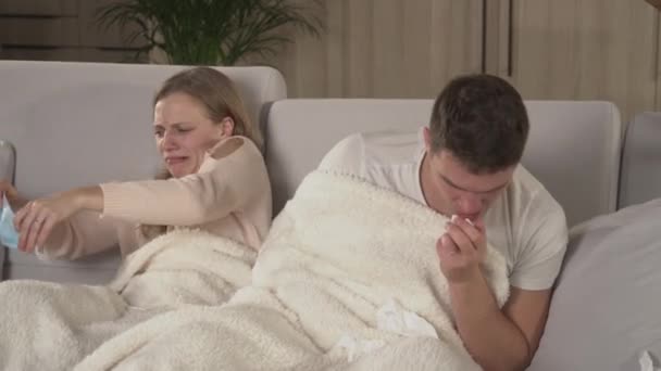 CLOSE UP: Young man sneezes and then young woman puts on a protective face mask. Autumn colds and flu spreading around. Married couple cuddling on comfy couch while man is having a seasonal virus. - Footage, Video
