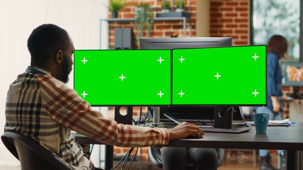 Hombre afroamericano analizando la pantalla verde en varios monitores, trabajando en un estudio de agencia creativa. Empleado masculino mirando la exhibición aislada con el copyspace de la llave del croma en la computadora. - Foto, Imagen