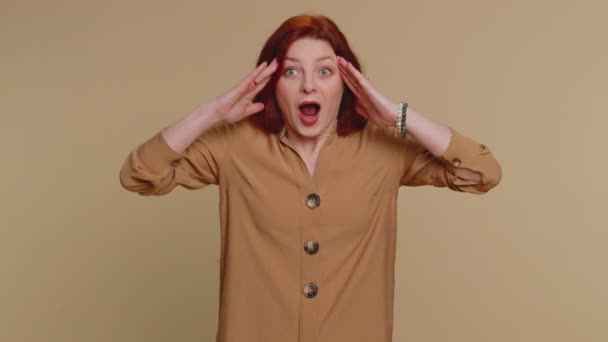 Excited amazed redhead woman in blouse touching head and showing explosion, looking worried and shocked, professional burnout. Looking surprised wow girl isolated alone on beige studio wall background - Footage, Video