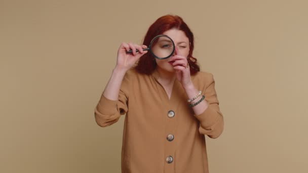 Onderzoeker onderzoeker wetenschapper vrouw houden vergrootglas in de buurt van gezicht, kijken in de camera met grote zoomde grappige ogen, zoeken, analyseren. jong roodharige meisje geïsoleerd op beige studio achtergrond - Video