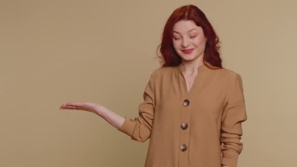 Lovely millennial redhead woman showing thumbs up and pointing empty place, advertising area for commercial text, copy space for goods promotion. Young girl. Studio shot indoors on beige background - Imágenes, Vídeo