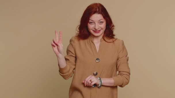 Хіпстерська руда жінка в блузці демонструє знак перемоги v, сподіваючись на успіх і перемогу, роблячи мирний жест, посміхаючись добрим оптимістичним виразом. Молода дівчина для дорослих ізольована на бежевому студійному фоні
 - Кадри, відео