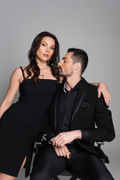 bearded man sitting on chair with sexy brunette woman in black dress isolated on grey  - Φωτογραφία, εικόνα