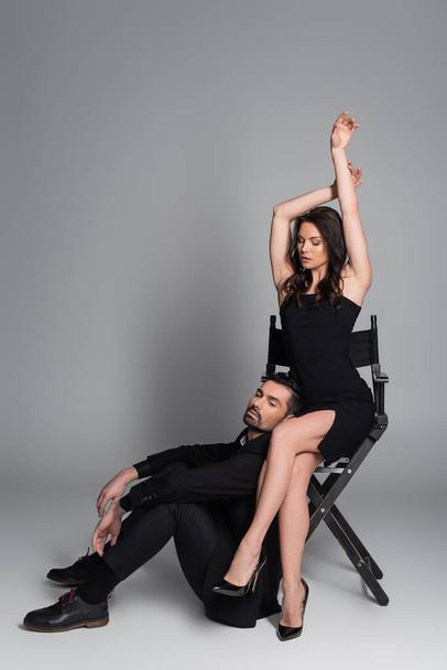 full length of brunette woman in black dress and high heels sitting on chair with raised hands near bearded man on grey  - Foto, Imagen