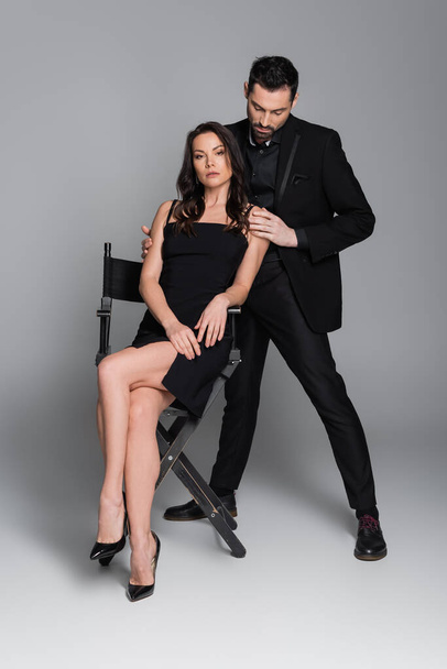 full length of brunette woman in dress and high heels sitting on chair near bearded man in suit on grey  - Foto, Imagen
