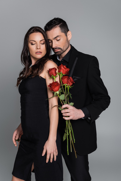 bearded man in suit holding red roses near brunette woman isolated on grey - Foto, afbeelding
