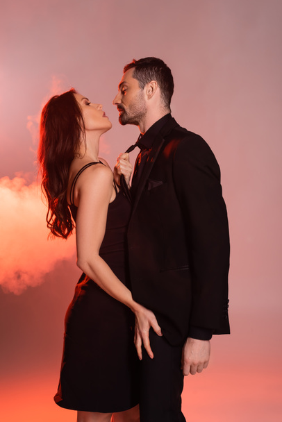 side view of sexy woman in black dress pulling tie while seducing confident man in formal wear on pink and grey  - Zdjęcie, obraz