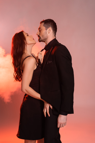 side view of sexy woman in black dress pulling tie while seducing bearded man in formal wear on pink and grey  - Foto, Imagen