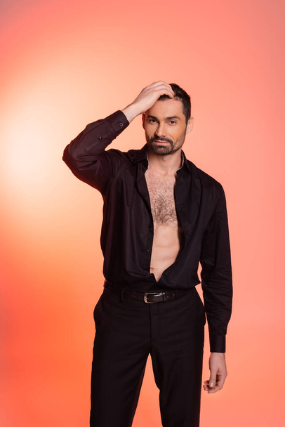 bearded man with hairy chest standing in black outfit and adjusting hair on pink  - Photo, image