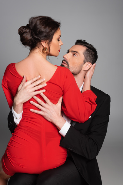 Vášnivý muž objímání sexy přítelkyně v červených šatech izolované na šedé  - Fotografie, Obrázek