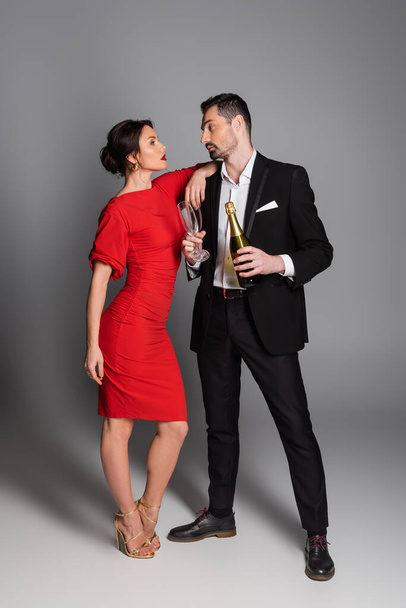 Элегантный мужчина в костюме держит бутылку шампанского и бокалы рядом с девушкой на сером фоне  - Фото, изображение