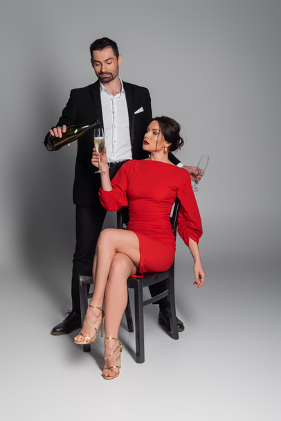 Elegante man gieten champagne in glas van vriendin in jurk zitten op stoel op grijze achtergrond  - Foto, afbeelding