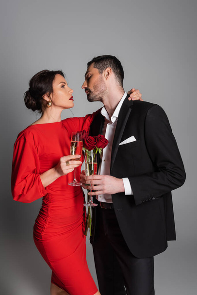 Side view of sexy woman in dress hugging boyfriend with champagne and roses on grey background  - Photo, Image