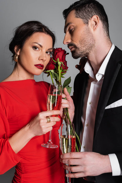 Elegantní žena dívá na kameru poblíž přítele se šampaňským a růží izolovaných na šedé  - Fotografie, Obrázek