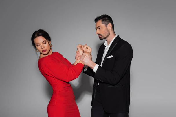 Elegante man houdt handen van de vrouw in rode jurk op grijze achtergrond  - Foto, afbeelding