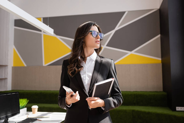 corporate manager in eyeglasses holding smartphone and notebook while looking away in office - Foto, imagen