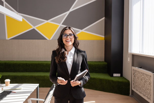 happy brunette businesswoman in eyeglasses looking at camera while standing with smartphone and notebook in office - Foto, afbeelding