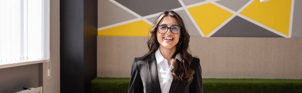 joyful businesswoman in eyeglasses and black blazer smiling at camera in office, banner - Foto, immagini
