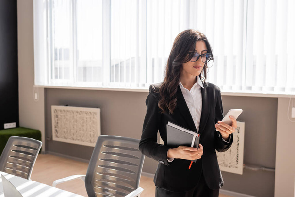 businesswoman in eyeglasses holding notebook and messaging on mobile phone in office - Photo, image