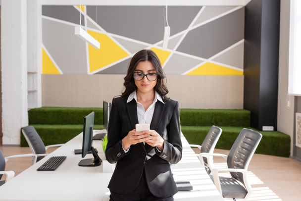 現代のオフィスのコンピュータと机の近くのスマートフォンで黒いスーツと眼鏡のメッセージングのビジネス女性 - 写真・画像