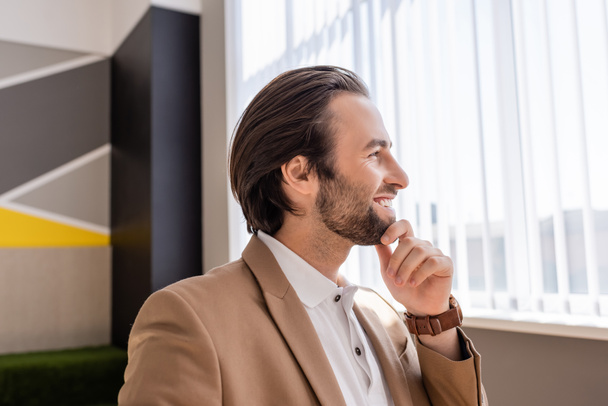 Улыбающийся бородатый бизнесмен трогает подбородок и смотрит в сторону в офисе - Фото, изображение