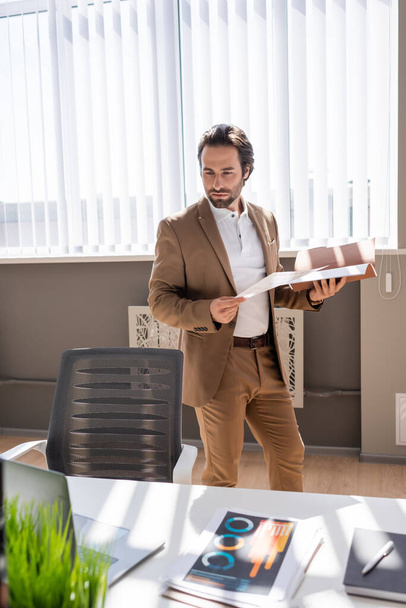 серьезный бизнесмен с документами во время просмотра ноутбука на столе в офисе - Фото, изображение