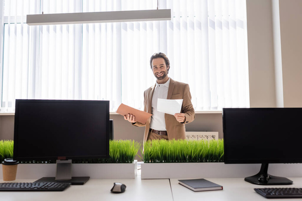 молодой бизнесмен с документами и улыбкой на камеру возле мониторов компьютера с чистым экраном в офисе - Фото, изображение