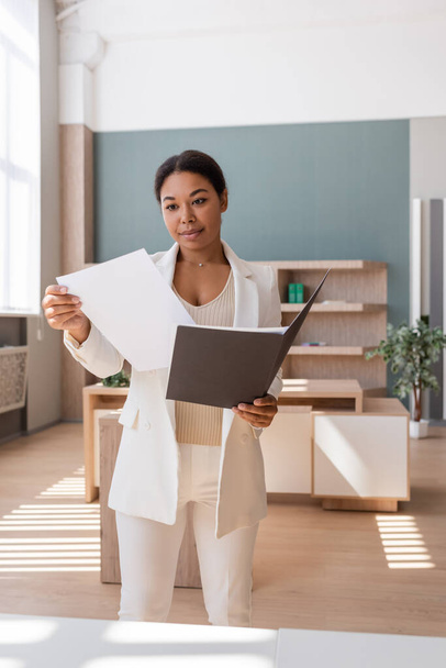 brunette multiracial woman in white blazer looking at documents in office - Fotó, kép
