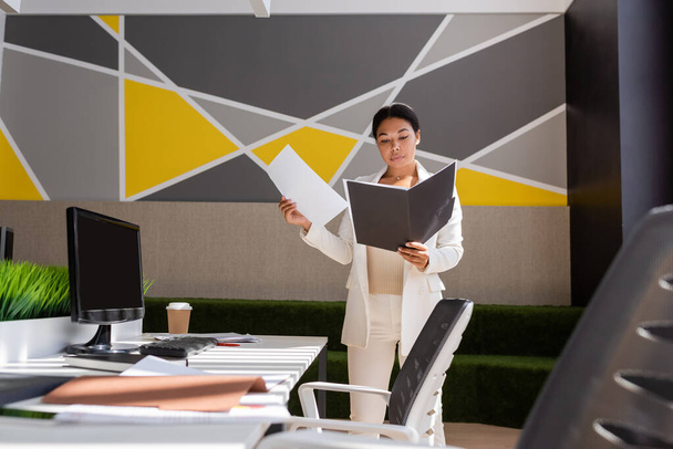 многорасовая деловая женщина, работающая с документами, стоя у стола в современном офисе - Фото, изображение