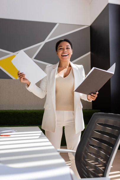 excited multiracial businesswoman in white suit holding documents and smiling at camera in office - Fotoğraf, Görsel